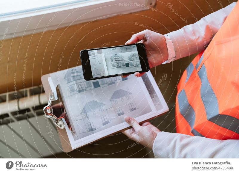 unrecognizable architect woman in construction site using mobile phone holding blueprints workspace protective helmet protective jacket real estate caucasian