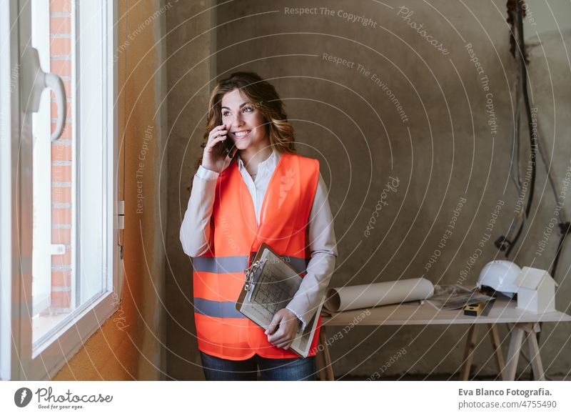 professional architect woman in construction site talking on mobile phone holding blueprints workspace protective helmet protective jacket real estate caucasian