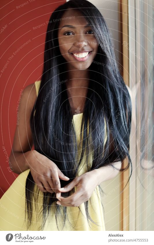 Young beautiful long haired woman in yellow summer dress stands in front of red wall at window and smiles at camera Woman Young woman pretty Beauty & Beauty