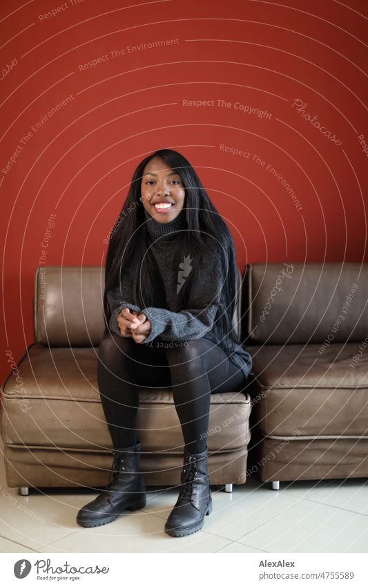 Young beautiful long haired woman sitting on brown leather couch in front of red wall and smiling at camera Woman Young woman pretty Beauty & Beauty Graceful