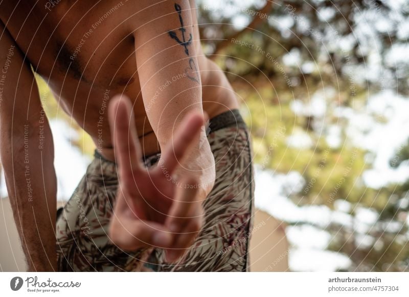 Young man in swimming trunks shows peace sign with fingers in shadow of tree youthful Man Peace Peace Symbols men masculinity Heart heart-shaped Fingers