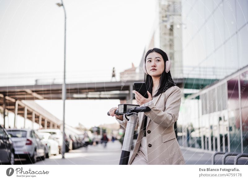 chinese business woman renting electric scooter in city,wearing headphones, using mobile phone music urban modern sustainability public transport asian young