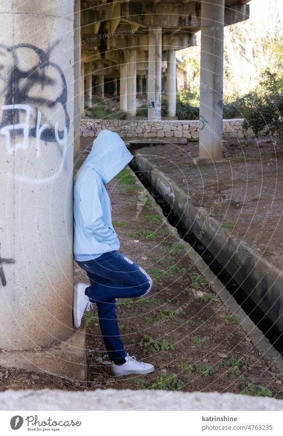 Hooded Teenage girl in light blue oversize hoodie stays under bridge Teenager adolescence jeans Blue teen girl outdoors bridge pillars graffiti urban modern