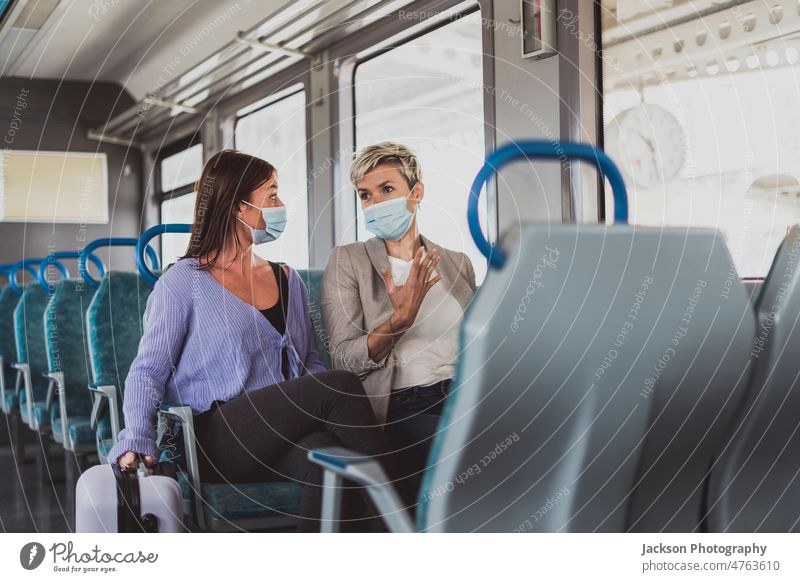 A couple of friends wearing mask and talking while traveling by train woman conversing chat women female bag luggage smartphone sitting new normal vacation