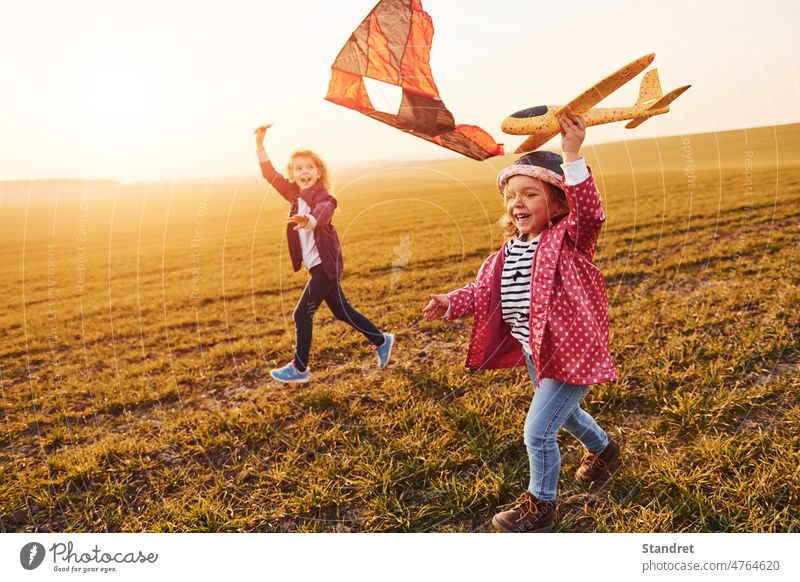 Two little girls friends have fun together with kite and toy plane on the field at sunny daytime unity kids children play run outside nature spring sunlight