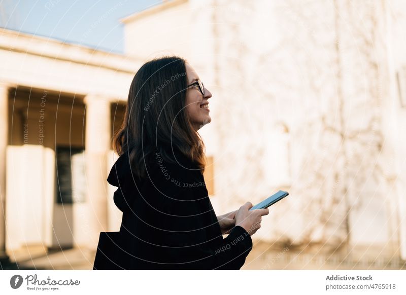 Smiling woman in eyeglasses and black hoodie using smartphone outdoors positive connection message millennial browsing city online female smile urban cellphone