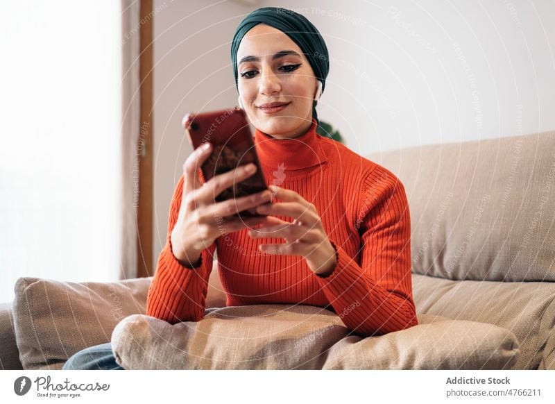 Muslim female using smartphone on sofa woman portrait home rest earphones earbuds tws wireless listen music social media weekend muslim ethnic islam playlist