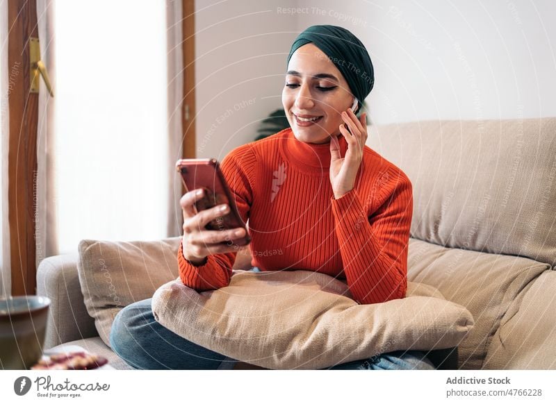 Muslim female using smartphone on sofa woman portrait home rest earphones earbuds tws wireless listen music social media weekend muslim ethnic islam playlist