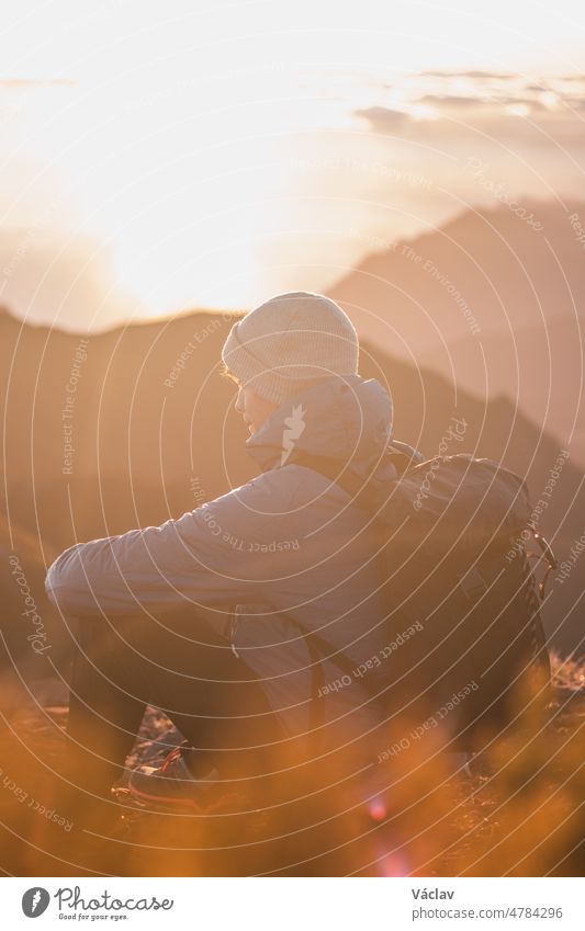 Young adventurer sitting on the edge of a rock on the highest mountain Pico Ruivo on the island of Madeira, Portugal at sunrise. The orange-gold light illuminates the young man. Candid portrait