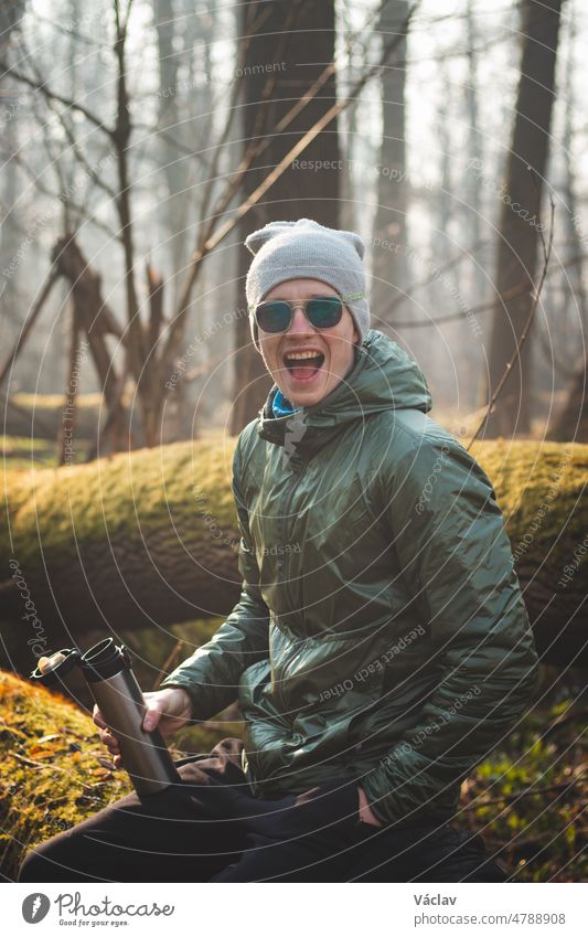 Laughing, experienced hiker sitting on a tree trunk in the wilderness, drinking his hot tea from a thermos. Real people discovering the biodiversity of Czech forests. A real smile