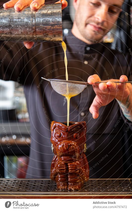 Crop barman pouring cocktail through strainer into tiki glass in bar drink prepare bartender barkeeper work shaker service counter male young uniform job