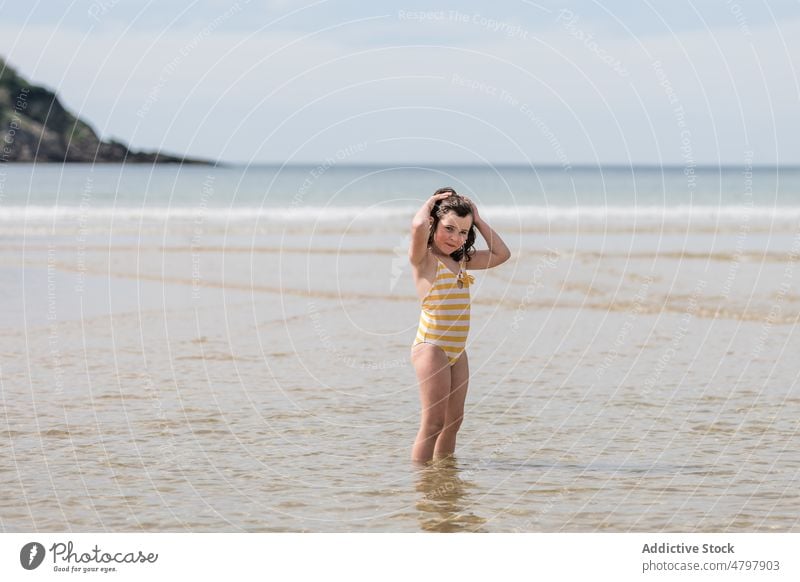 Girl standing near sea looking at camera kid girl beach coast childhood rest shore water recreation wave wind flying hair swimsuit swimwear leisure sand