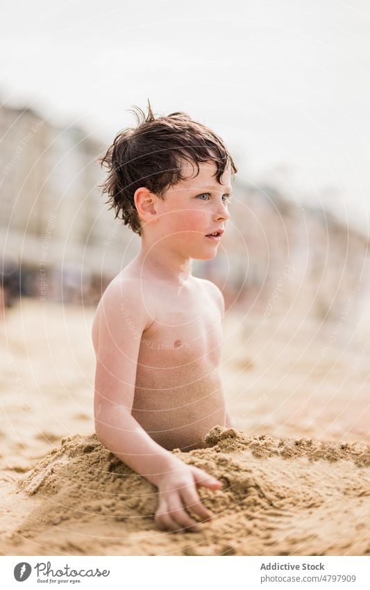 Boy burying himself in sand looking away boy kid play beach playful carefree game coast summer childhood rest shore pastime recreation leisure seashore seacoast