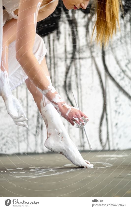 Portrait in profile of a young woman in white ballet dress holding a brush and paints her feet with paint in front of black and white artistic background. Woman