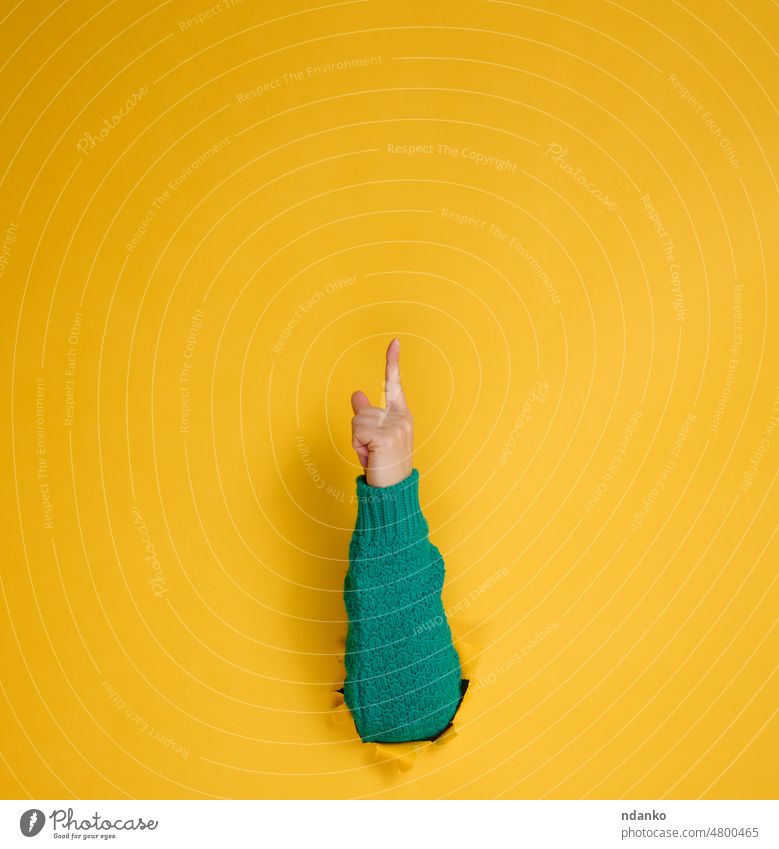 female hand sticks out of a torn hole in a yellow paper background, part of the body points with the index finger to top. Copy space adult arm attention break