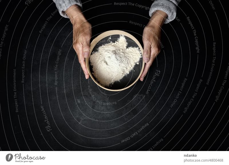 two female hands hold a round wooden sieve with wheat flour on a black table, top view food ingredient preparation cooking baking sifting kitchen white dough