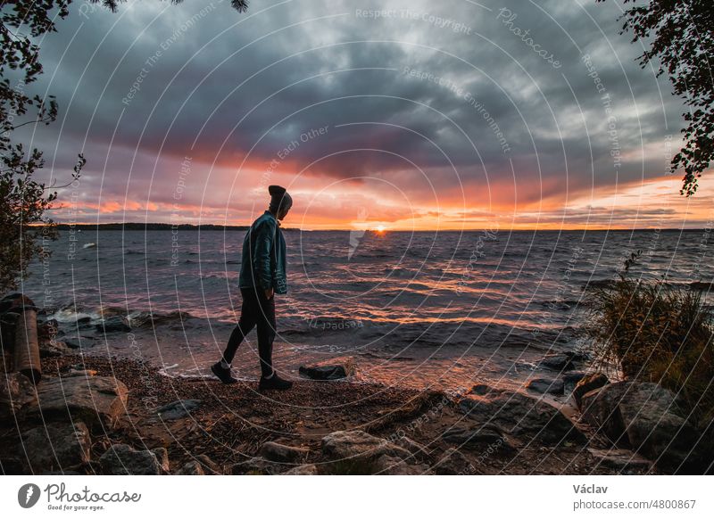 Dramatic sky with a mix of red, orange and purple over the waves of Lake Oulujarvi with a view of a man in a blue shirt and cap watching this gem on the beach at Kajaani, Kainuu region, Finland