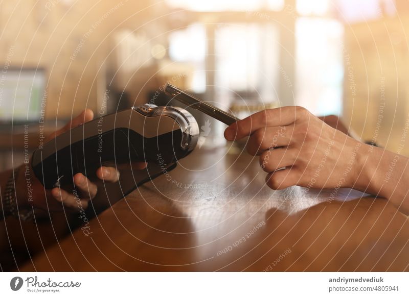 Macro unknown hand giving terminal to visitor for mobile phone payment indoor. Client hand paying bill with smartphone in blurred background. Cropped shot hands making contactless transaction inside.