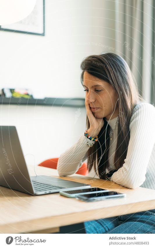 Focused young ethnic woman sitting at table with laptop and smartphones freelance thoughtful online hand on cheek work connection remote watch analyze focus