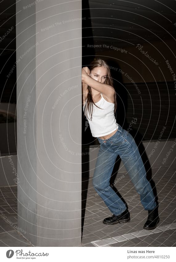 Parking lot with a gorgeous brunette girl standing next to a column. Dressed in dirty blue jeans and a white top she has this casual city life vibe. With kick-ass raw fashion looks.