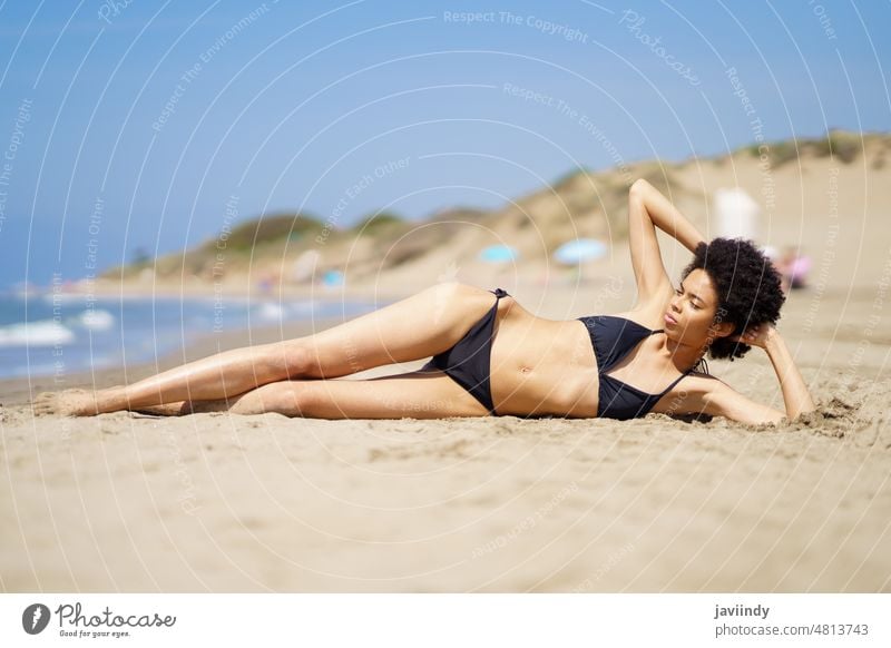 Relaxed black woman lying tanning on the sand of a tropical beach sunbathing. bikini summer swimsuit afro hairstyle relax swimwear coast curly background