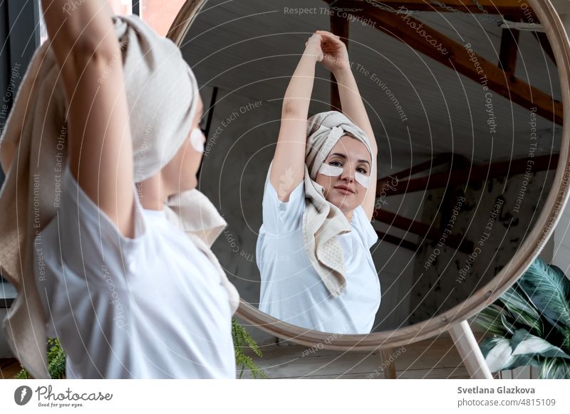 Young girl with towel on head, eye patches, looks mirror, make facial mask, takes care of beauty and mental health, skin care treatment, spa day relax wellbeing