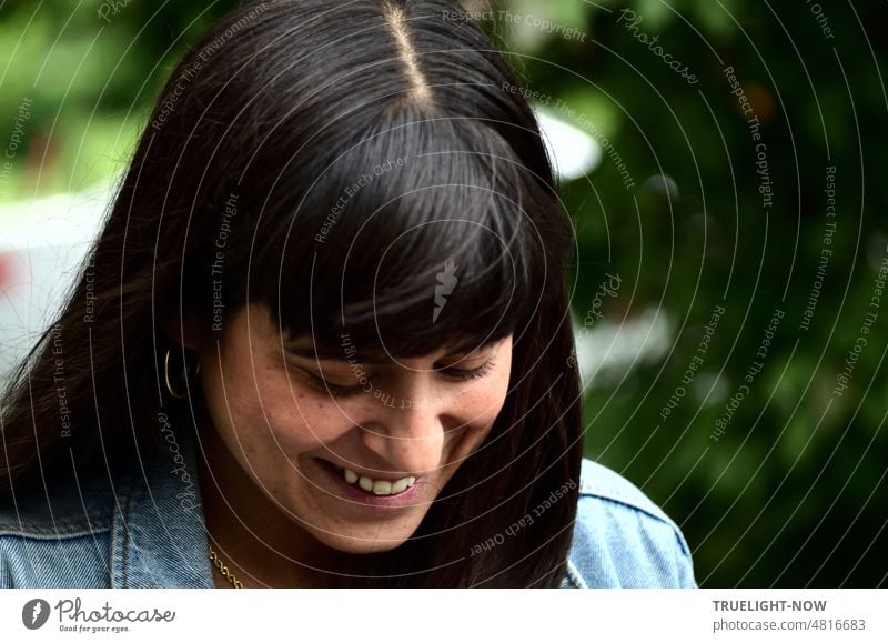 [Hansa BER 2022] Cheerful young woman outdoor close with long black hair, looking down, laughing, showing accurate parting and bangs cut, shoulder part a washed out denim jacket and from dark hair flashes an earring