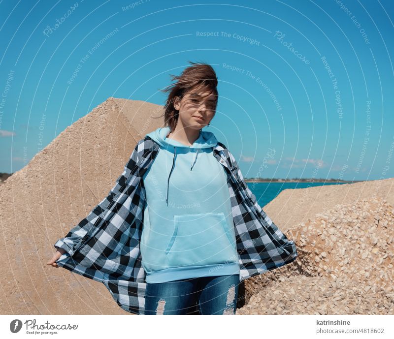 Teenager girl in light blue hoodie and plaid shirt against concrete blocks and blue sky mockup Blue short haired teen girl outdoors windy smiling hipster