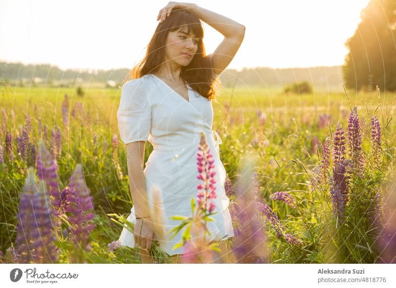 Beautiful woman in white sundress enjoying the summer nature. Picking colourful flowers, breathing fresh air and floral scent, walking in the sunny field of lupins. Happiness concept