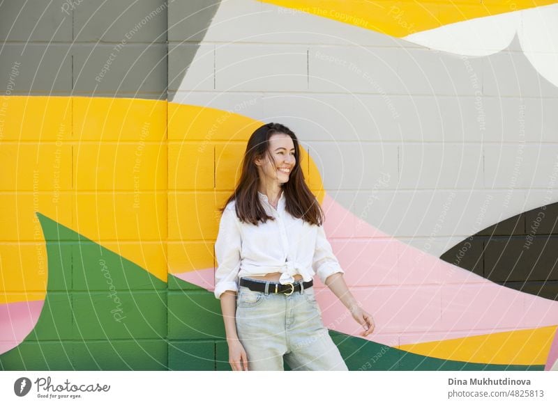 Young woman in white shirt and light blue jeans standing near colorful vibrant wall. Candid lifestyle summer style portrait. beautiful city happy urban girl