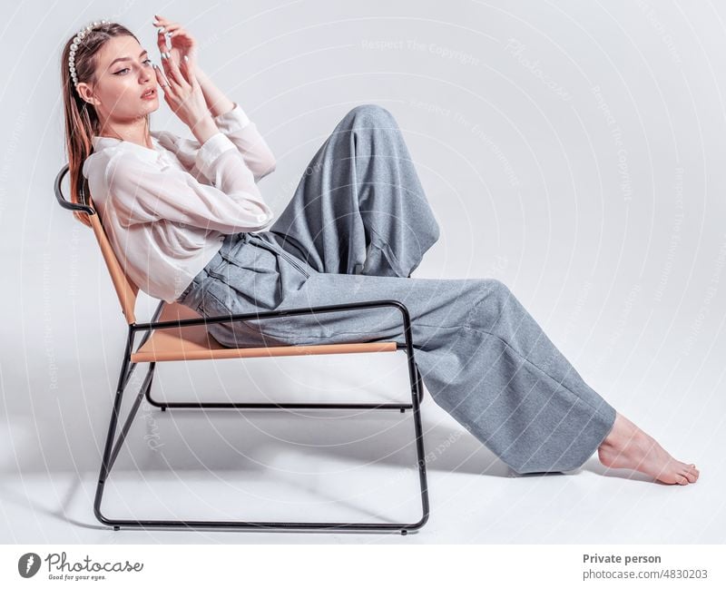 A beautiful young woman full height in pants and a white shirt sits barefoot in a chair against the background of the studio. Women sit-in Only one woman