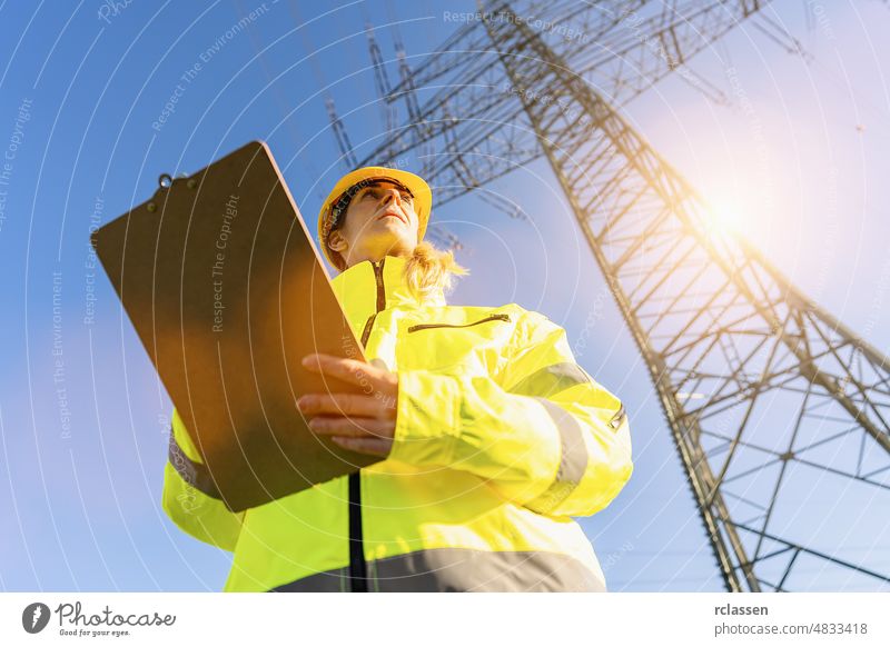 female electrical engineer standing with clipboard and watching at the electric power station to view the planning work by producing electricity at high voltage electricity poles.