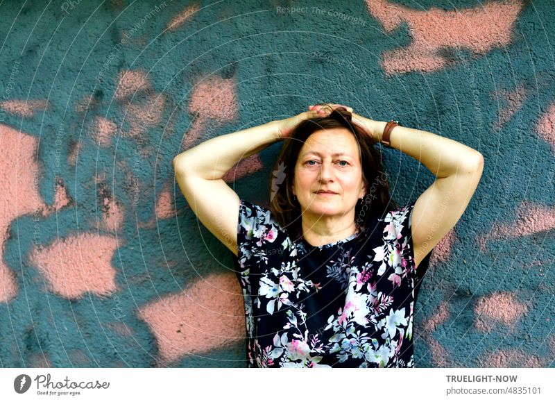 A lady in front of large pink blue graffiti in contrasting matching flowered summer dress with naked arms folded over head and wristwatch on wrist carefully shows her muscles smiling into camera