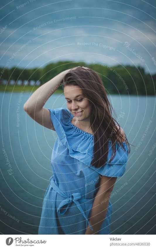 Cute brunette model with long straight dark hair in a summer blue dress with a beautiful realistic smile poses by a lake in the Czech Republic. European woman. Candid portrait with realistic smile