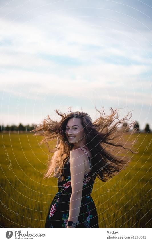 Breathtaking brunette model with a natural wide smile in a dark summer floral dress walks through the tall grass, her wavy dark hair flying. Candid portrait real adult woman. Czech republic, Europe