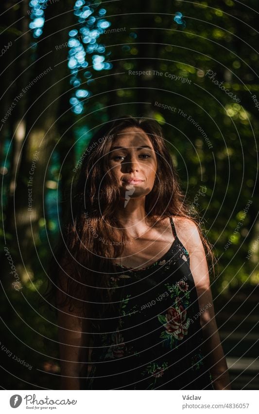 pensive view of a 24 year old woman with dark brown hair in a forest at sunset. Candid portrait of handsome woman in summer dress. Hidden in the shadows