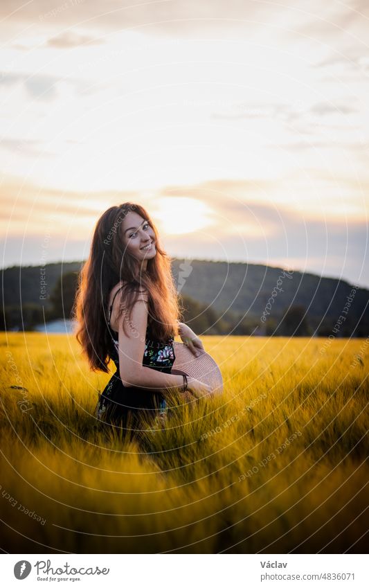 Beautiful realistic smile of brunette in black dress standing in the middle of a field during sunset. Candid portrait of inner emotions. Ruffle your hair summer