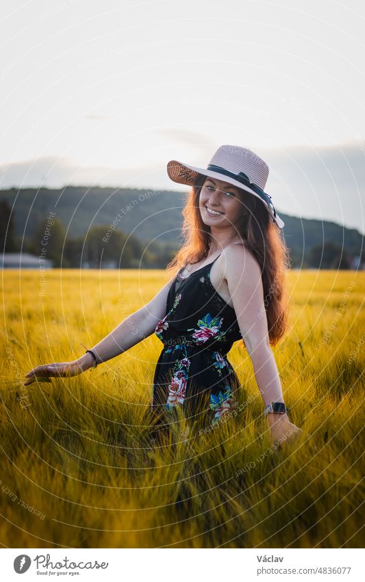 Wonderful natural smile of a full grown lady in a flowered dress with brown hair in the middle of a meadow wearing a beautiful wicker hat. Candid portrait of young model at sunset