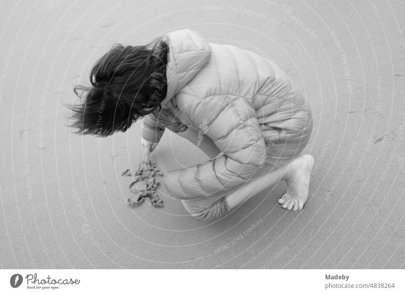 A boy with long hair and a thick down jacket writes something in the sand on the beach in Knokke-Heist on the North Sea near Bruges in West Flanders in Belgium, photographed in classic black and white