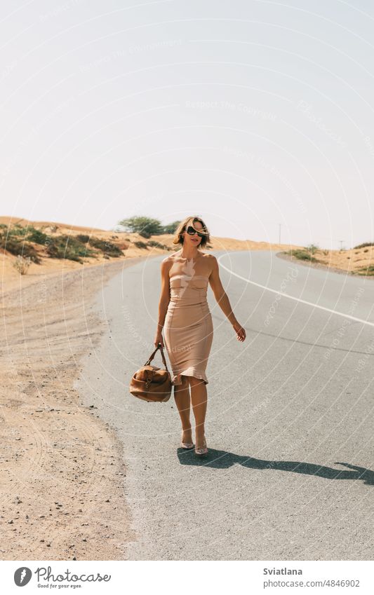 A glamorous girl in an elegant dress walks along a deserted road with a bag in her hand. glamour style beautiful woman portrait blond fashion beauty model young