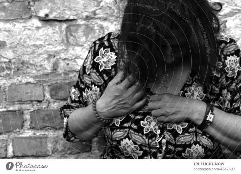 Lady in flowery summer dress is standing by a brick wall, her head is lowered to her chest so that her long dark hair hides her face and her hands seem to be intensely busy at the neckline of her dress