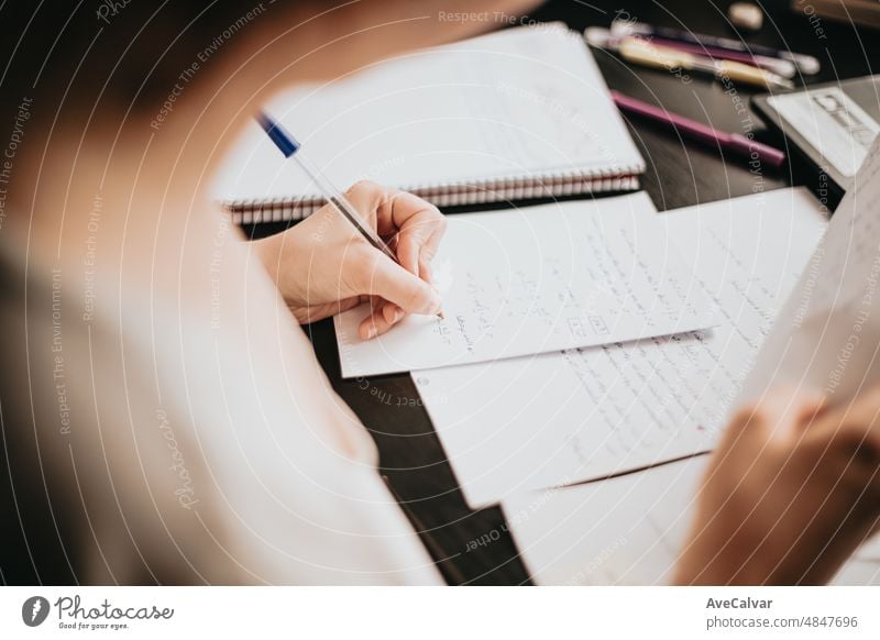 Close up detail image of a young woman studying and working on his home desk, doing homework during university, preparing for exam with textbook and taking notes, selective focus on the pencil.