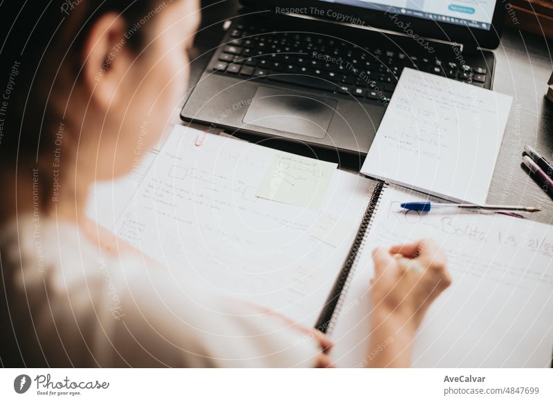 Close up detail image back of a young woman studying and working on his home desk, doing homework during university, preparing for exam with textbook and taking notes, selective focus on the pencil.