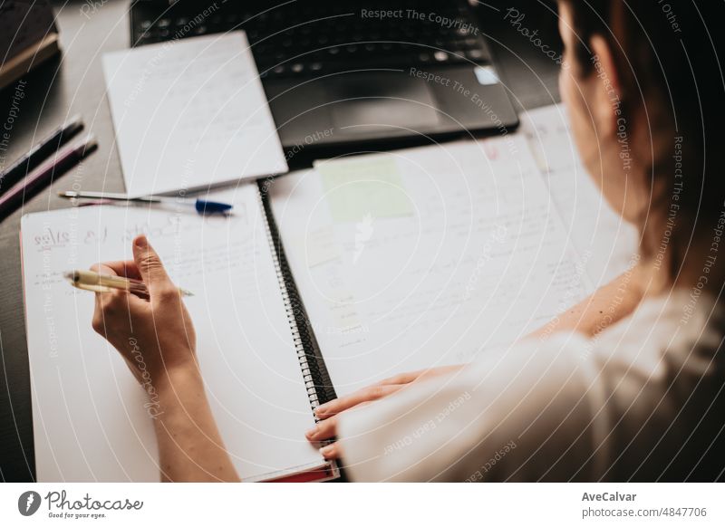 Top view of the back of a young woman studying and working on his home desk, doing homework during university, preparing for exam with textbook and taking notes, selective focus on the pencil.