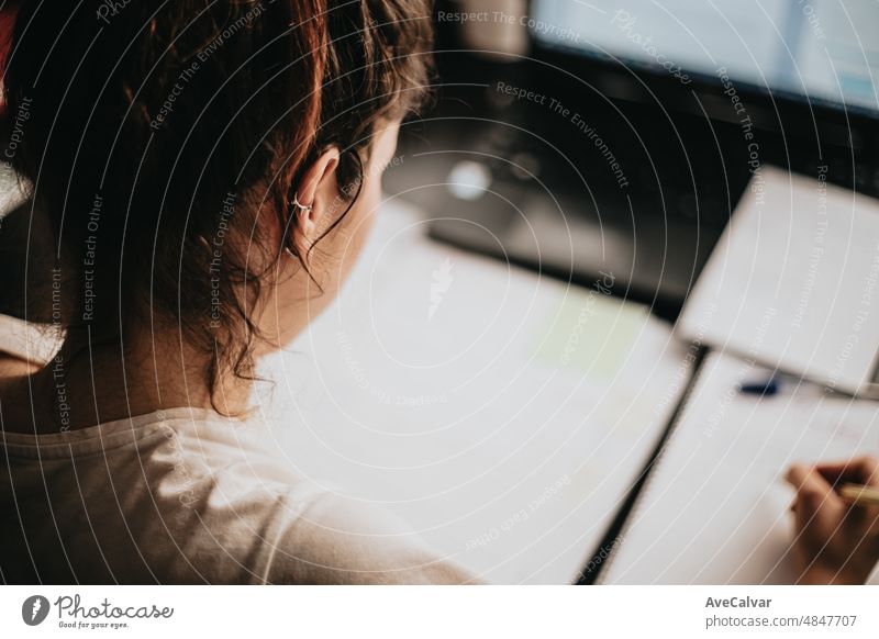 Close up detail image of a young woman studying and working on his home desk, doing homework during university, preparing for exam with textbook and taking notes, selective focus on the pencil.