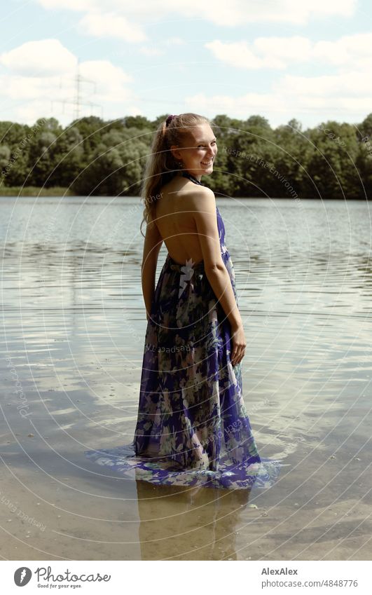 Young red blonde long haired woman stands in dress with feet in lake on shore and smiles sideways at camera Woman youthful pretty Slim Grass Nature Blonde