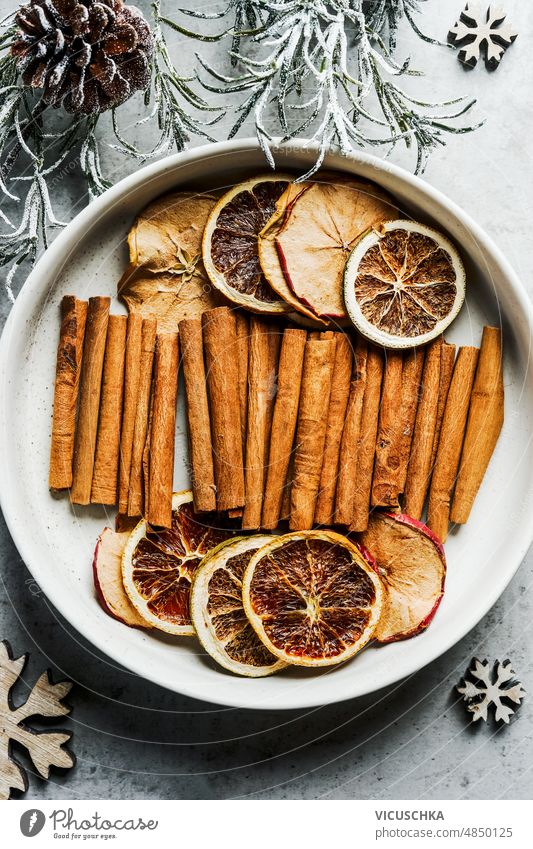 Dried citrus slices and cinnamon sticks in white bowl with fir green and pine cones. Winter flavor dried winter fruits spices top view background dried orange