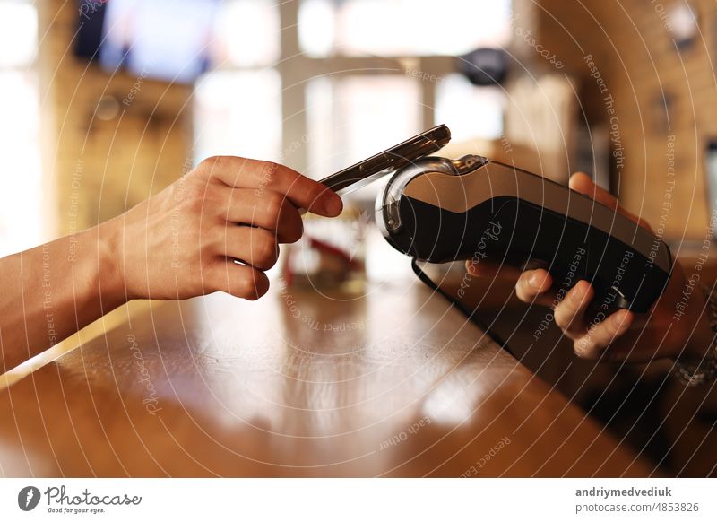 Macro unknown hand giving terminal to visitor for mobile phone payment indoor. Client hand paying bill with smartphone in blurred background. Cropped shot hands making contactless transaction inside.