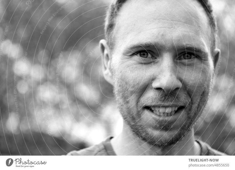 Black and white frontal portrait of young man and father, fearless smiling intense look into camera, short hair, 3 day beard, sunlight reflections out of focus in background