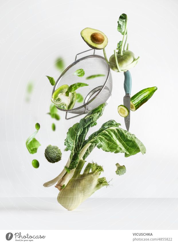 Balancing flying green vegetables ingredients: pak choi, avocado, fennel, bell pepper broccoli, edamame, turnip cabbage with sieve and kitchen knife at white background.